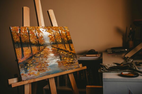 An oil painting of a fall landscape with vivid colors displayed in an artist's studio on a wooden easel.